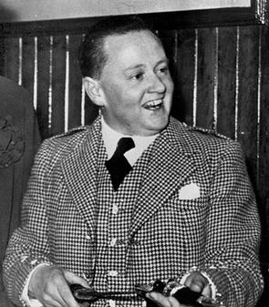A black and white photograph of a smiling white man, wearing a chequered suit.