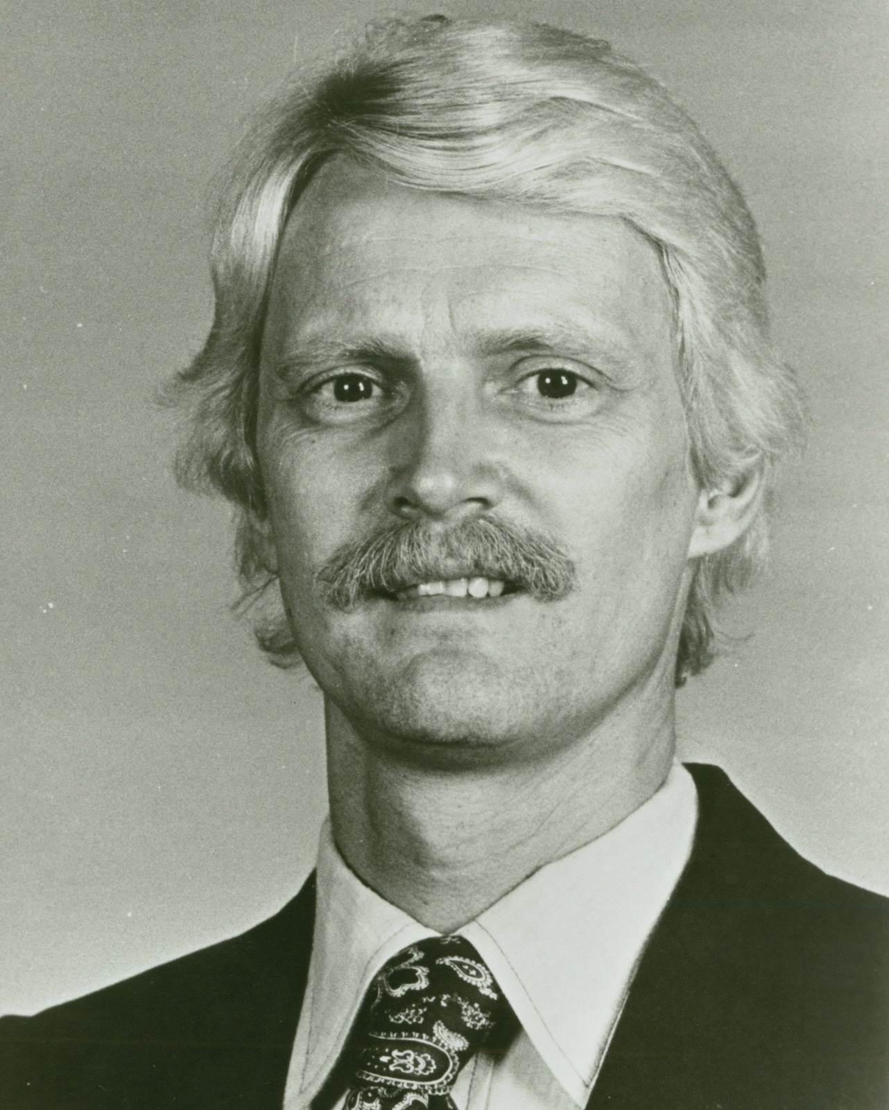 A black and white photograph of a white man with blonde hair and a mustache wearing a suit and paisley tie.